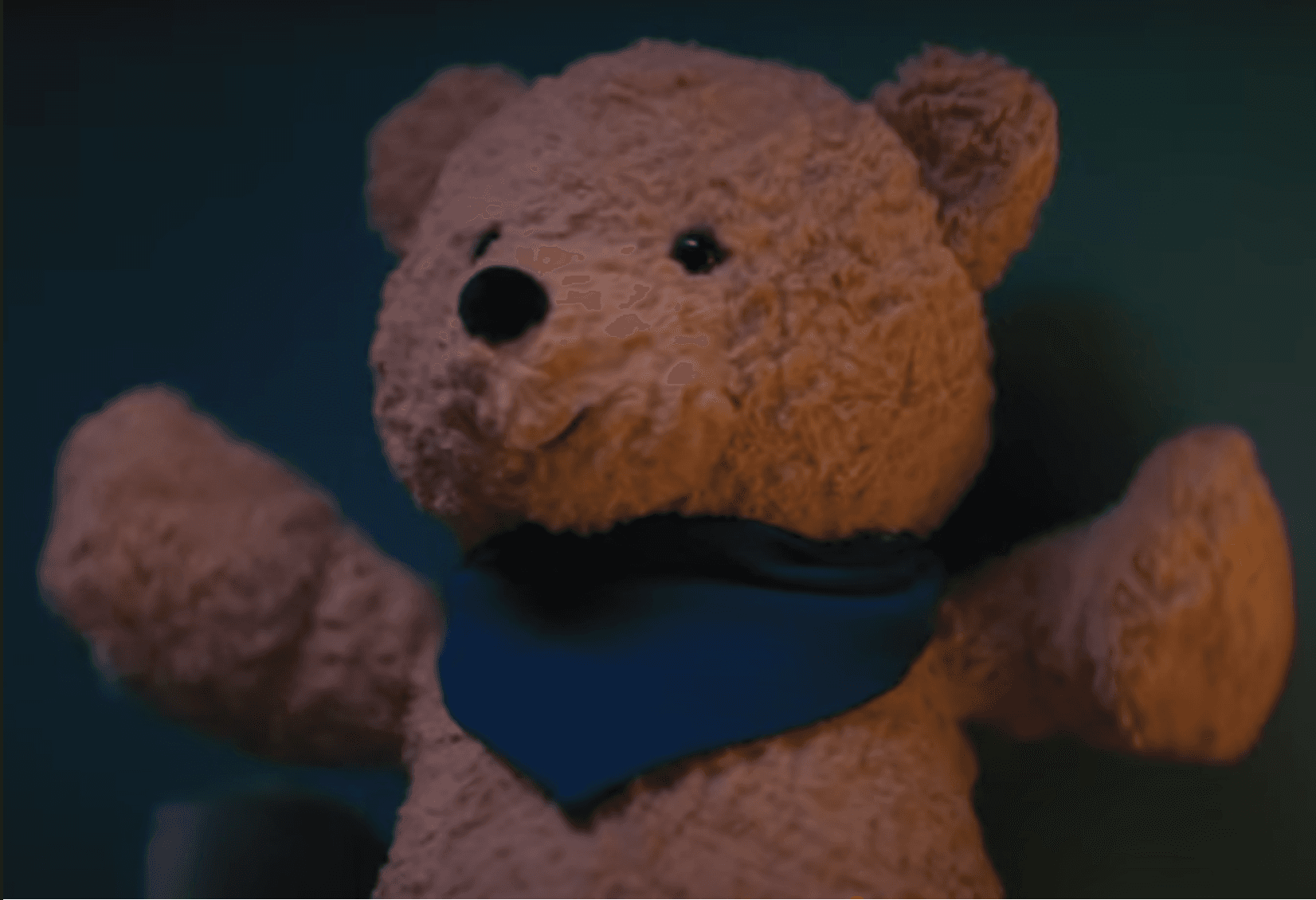 Fuzzy brown teddy bear wearing a blue bandana with arms outstretched against a teal background.