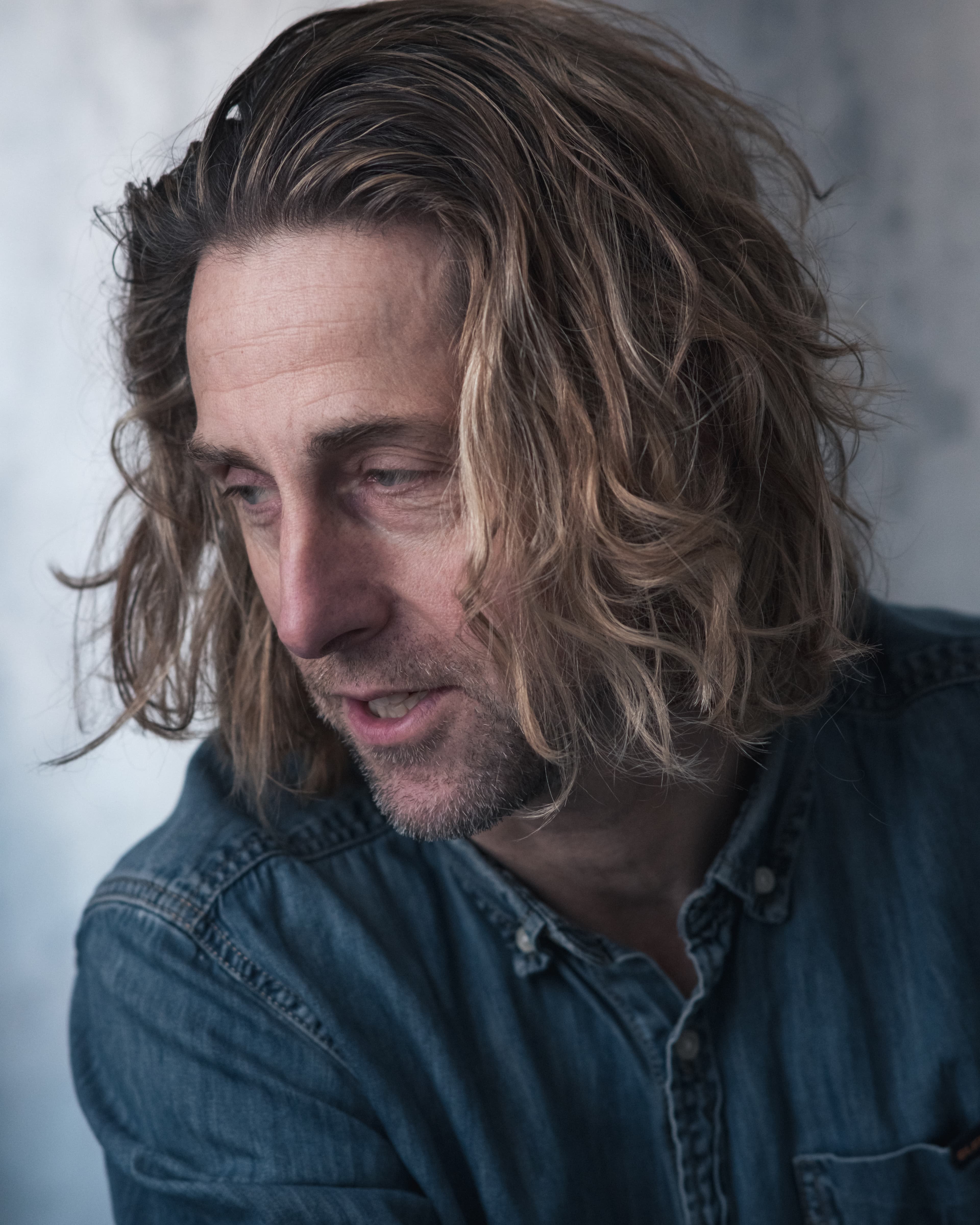 Close-up portrait of a man with long wavy hair and stubble wearing a denim shirt.