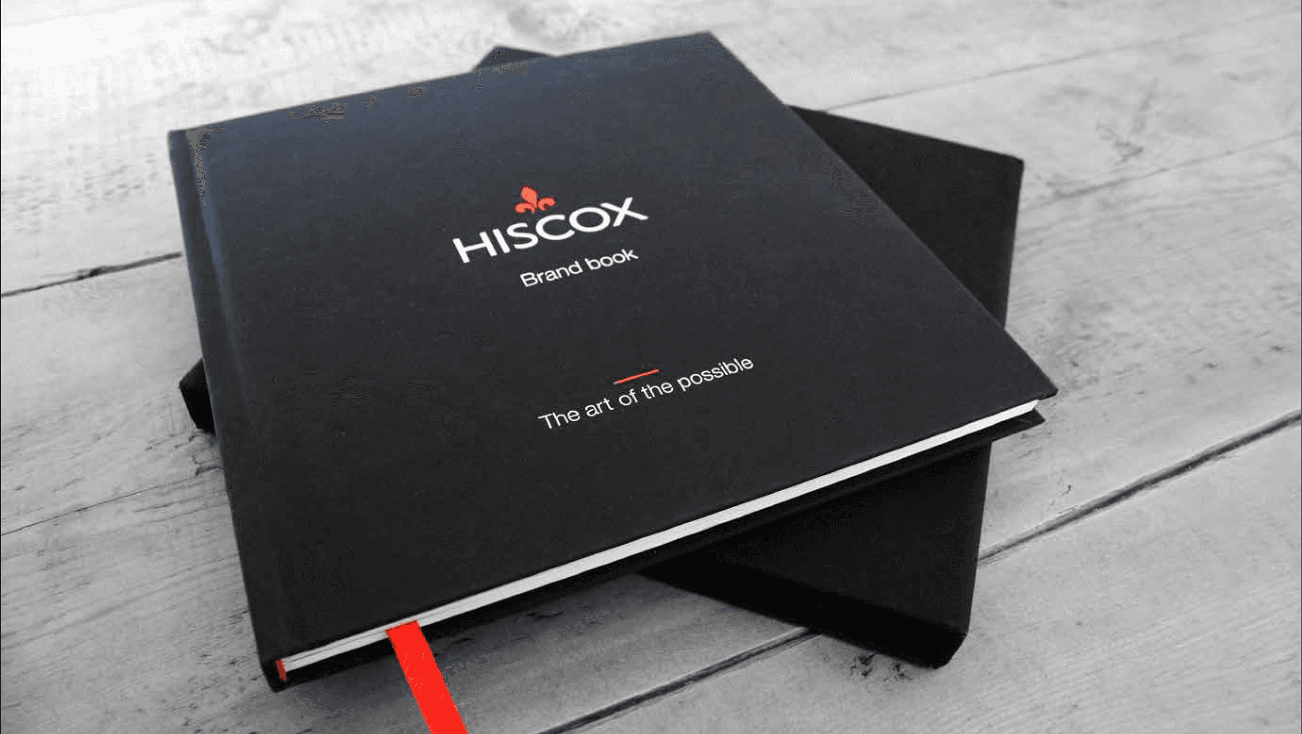 Two black Hiscox Brand books with red ribbon bookmarks stacked on a light wooden surface.